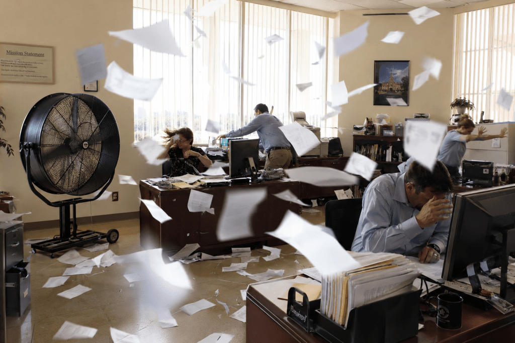 Created by fogBlogger using the ChatGPT prompt "I need a photorealistic image of a university administration office containing several people working at their desks. The day should be very sunny and bright and also extremely warm. In one corner of the room, there should be a giant, stand-up cooling fan running. The fan should be so strong that papers are flying around the office in a giant windstorm." In the public domain.