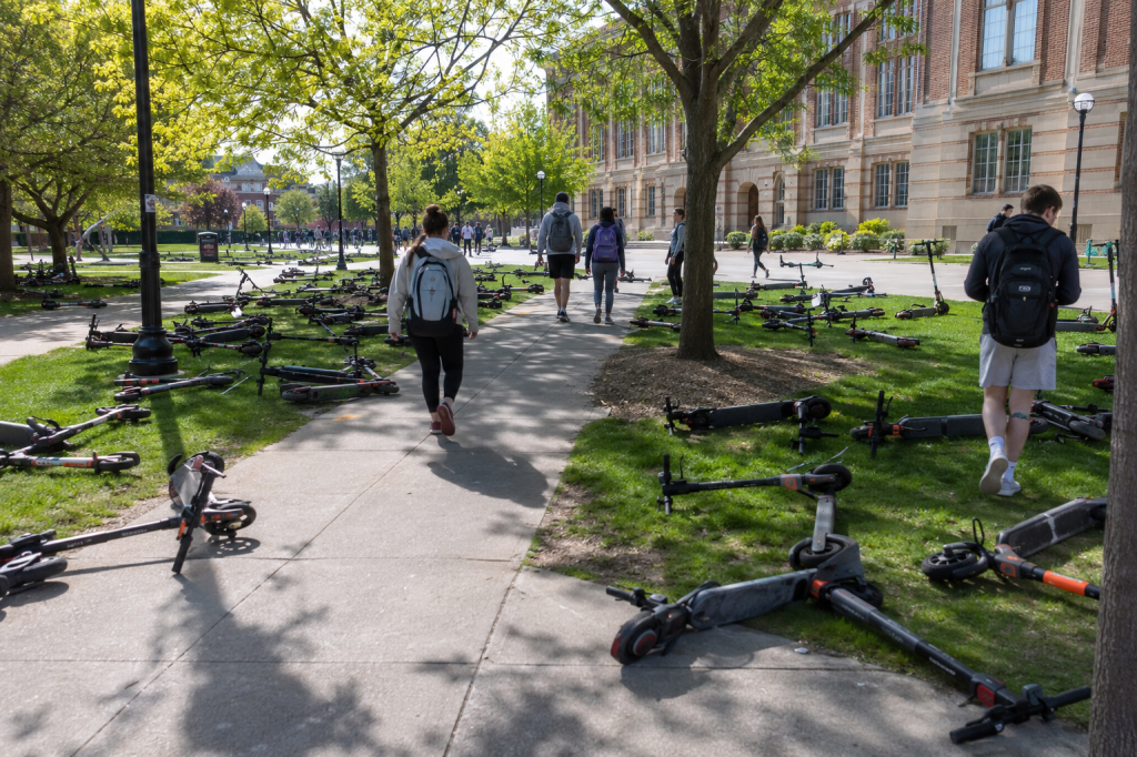 Created by ChatGPT using the prompt "I need a photorealistic image of a university campus. The day should be sunny and bright. Electric scooters should be lying haphazardly and in great numbers on all the sidewalks and grassy areas, forcing pedestrians to have to walk around them." In the public domain.